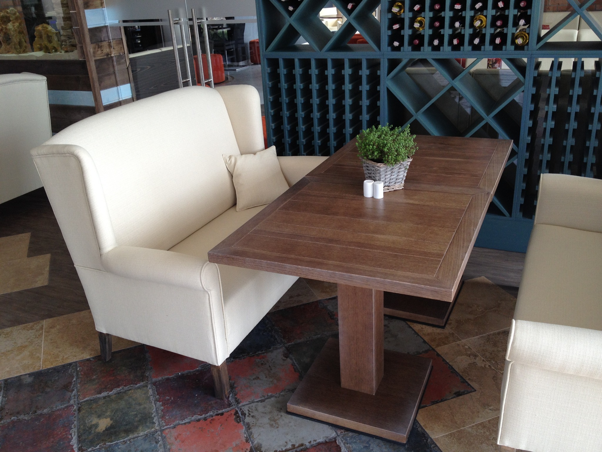 Nowoczesna sofa z wysokim oparciem w restauracji La Riva, obok drewniany stół z dekoracją roślinną i regał na wino w tle. Projekt Joanna Kukulska.