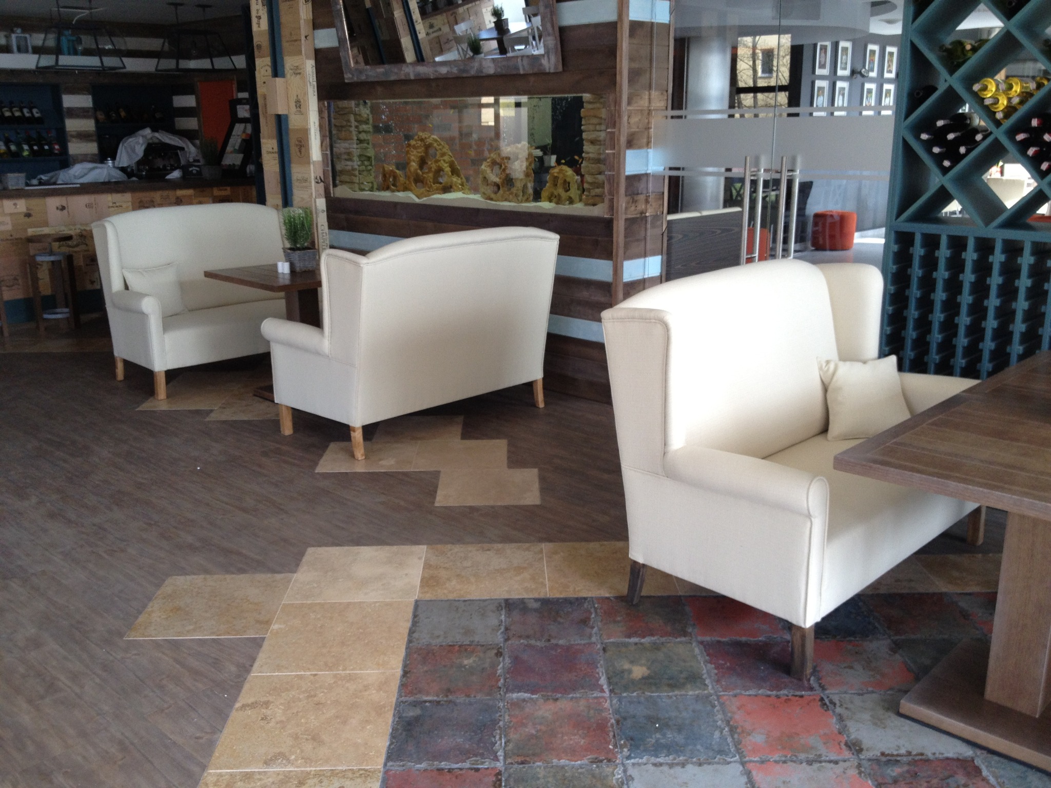 Eleganckie, kremowe sofy z poduszkami i drewnianymi stolikami w restauracji z akwarium w tle. Nietypowa podłoga z płytek w różnych kolorach i wzorach.