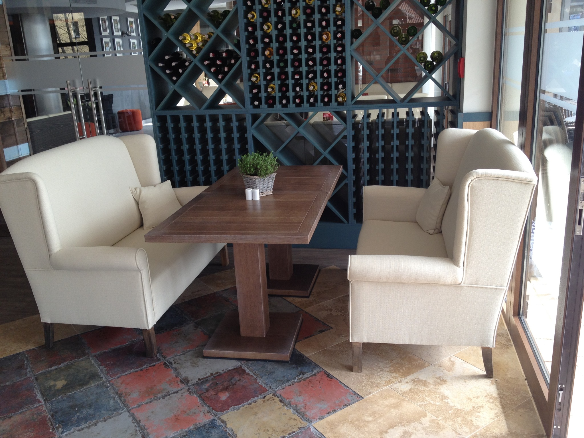 Beżowe fotele klubowe z drewnianym stolikiem i dekoracją roślinną na tle regału na wino w restauracji. Widok na patchworkową podłogę i okno.