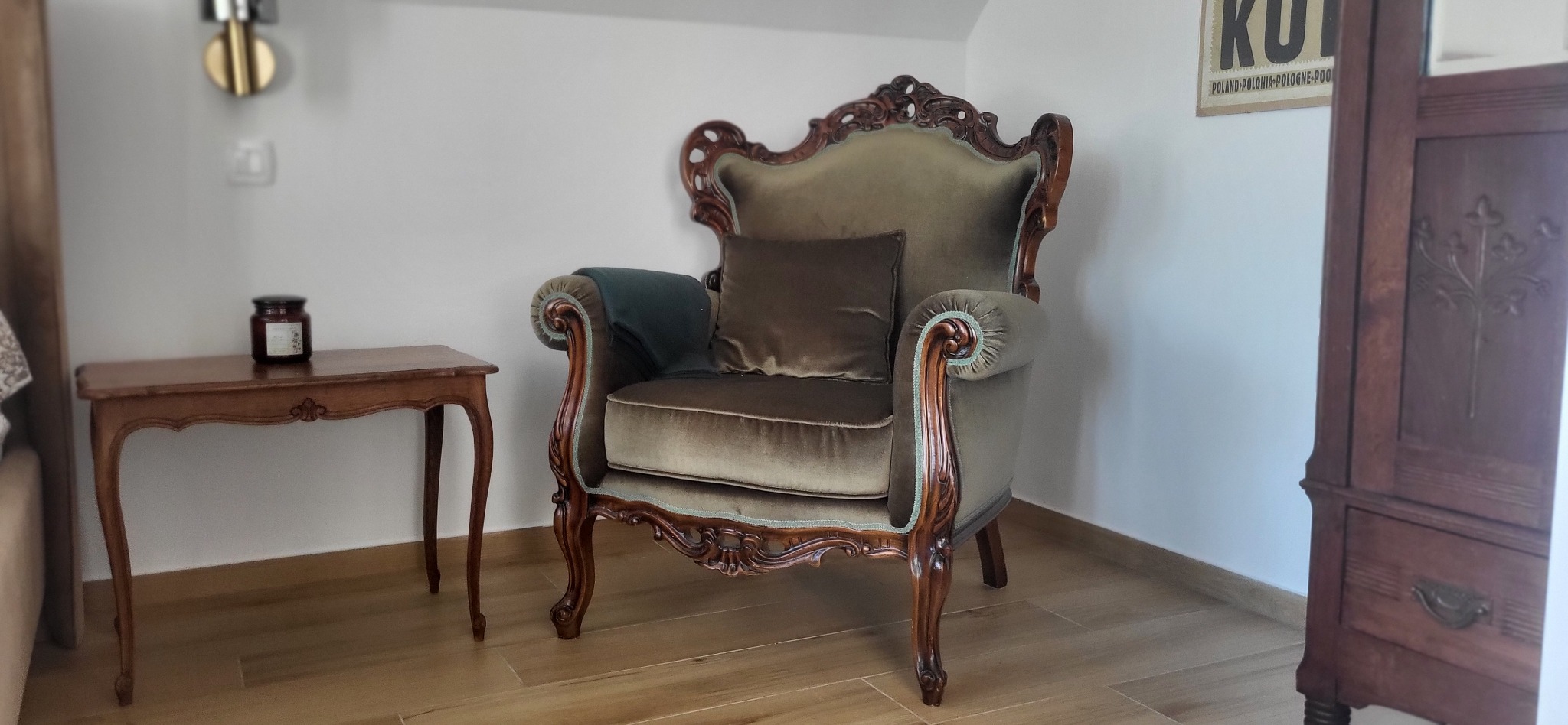 Elegancki fotel w stylu vintage, obity pluszowym welurem w odcieniach zieleni i brązu, z rzeźbionymi drewnianymi elementami, na tle jasnego wnętrza z drewnianą podłogą.