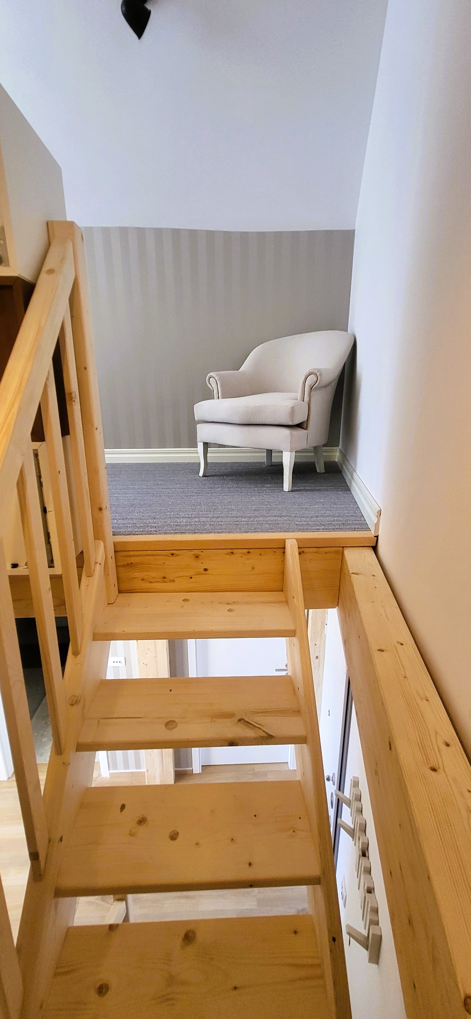 Kremowy fotel butikowy na tle szaro-białej ściany, widziany z perspektywy drewnianych schodów. Ujęcie z dołu, podkreślające unikalny kąt widzenia.