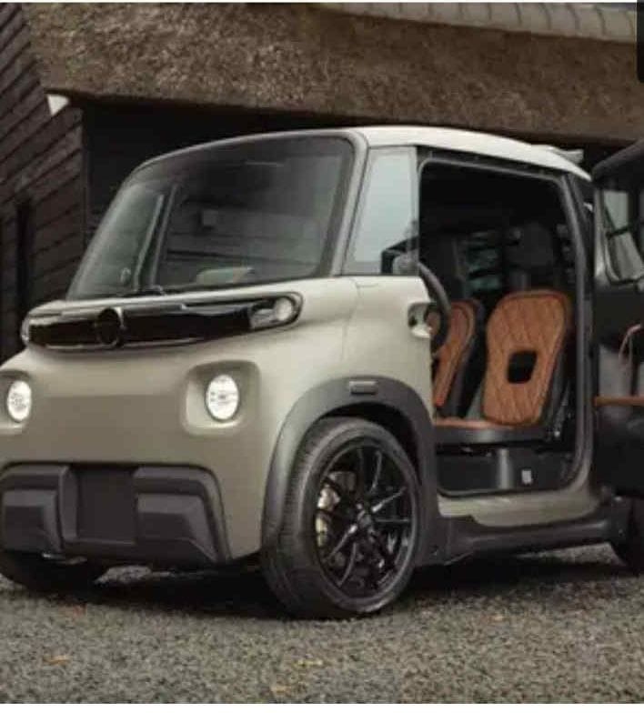 Beżowy, elektryczny mikrosamochód z otwartymi drzwiami, ukazujący brązowe, pikowane siedzenia. Czarne felgi i nowoczesny design pojazdu.