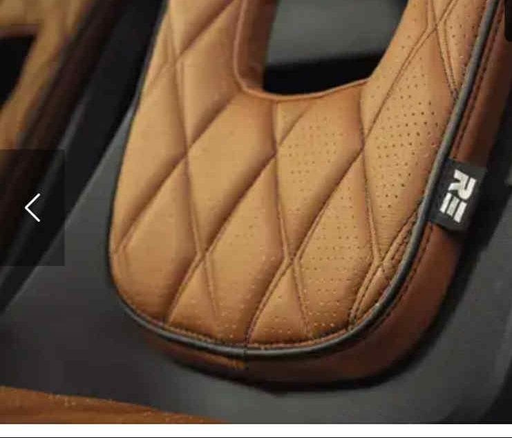 Brązowe, perforowane siedzisko z pikowaniem w romby, wykończone czarną lamówką i metką z logo, na ciemnym tle. Detal tapicerki samochodowej.