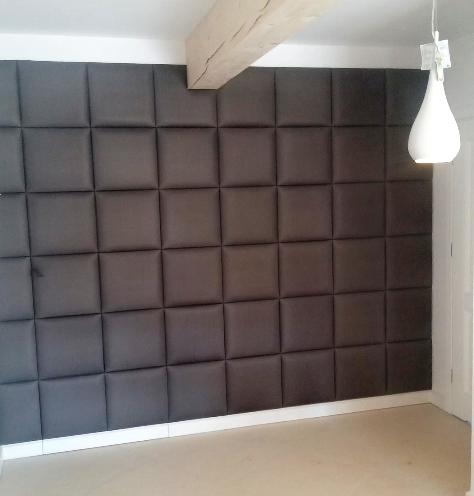 Tapicerowana ściana z ukrytym skrzydłem drzwiowym, wykończona w ciemnoszarych, kwadratowych panelach. Widoczna drewniana belka stropowa i minimalistyczna lampa.