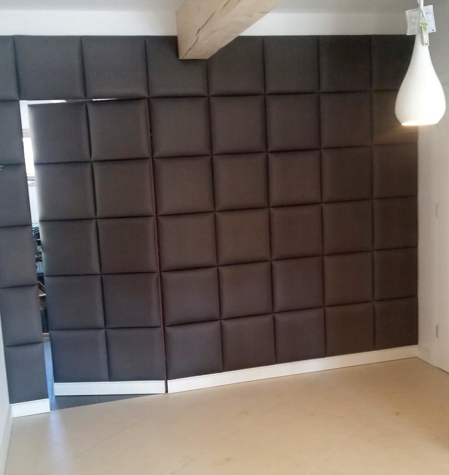 Ściana z brązowych, tapicerowanych paneli w kształcie kwadratów, z widocznym fragmentem drewnianej belki stropowej i lampą. Nowoczesny design wnętrza.