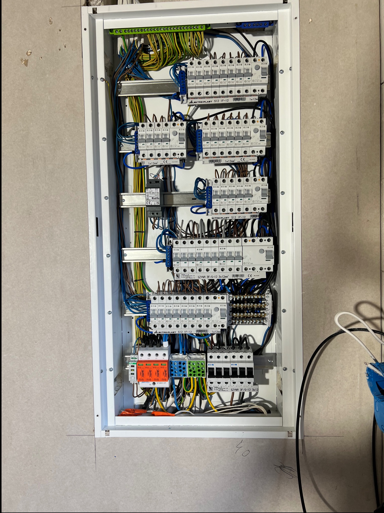 Rozdzielnica elektryczna wnękowa z zainstalowanymi bezpiecznikami, przewodami i listwami, widok z bliska na szarej ścianie.