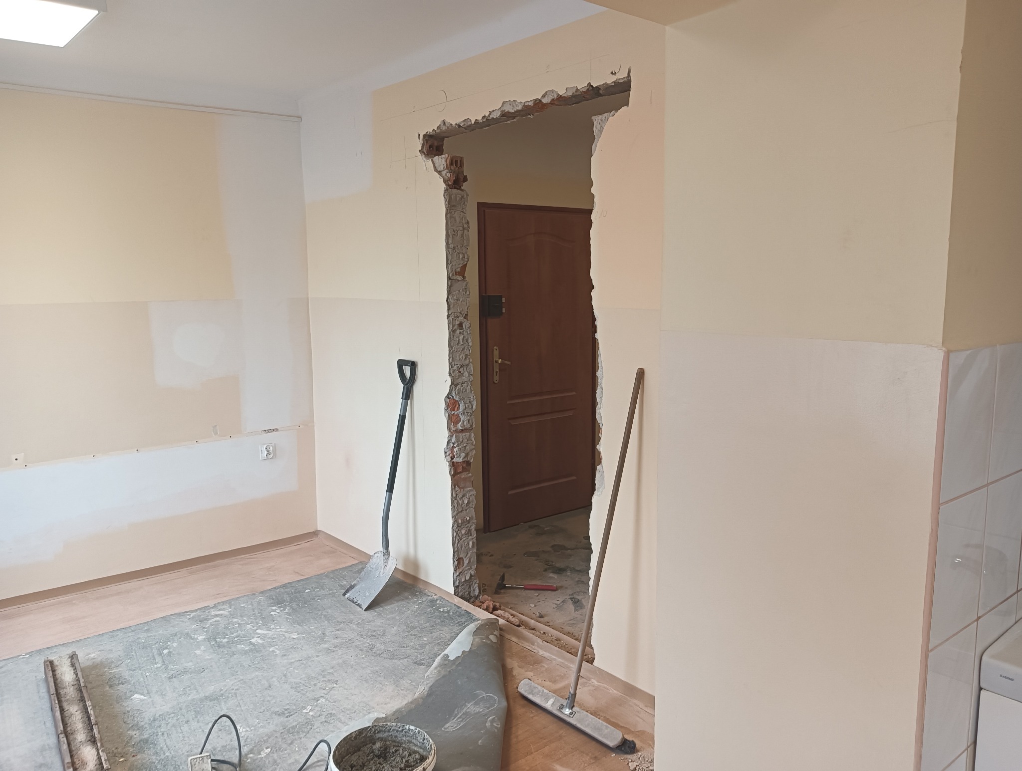 Wyburzona ściana działowa z widocznymi fragmentami cegieł, łopata i miotła oparta o ścianę, wiadro z zaprawą, podłoga w trakcie demontażu.