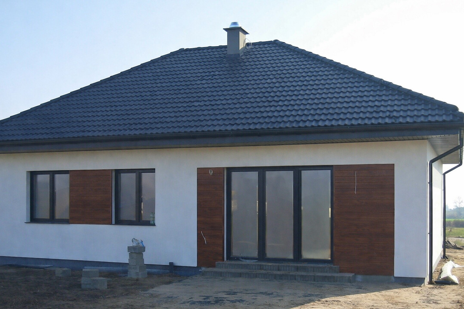 Parterowy dom z ciemną dachówką i drewnianymi panelami elewacyjnymi. Widoczne okna i przeszklone drzwi tarasowe. Na pierwszym planie ułożone bloczki.