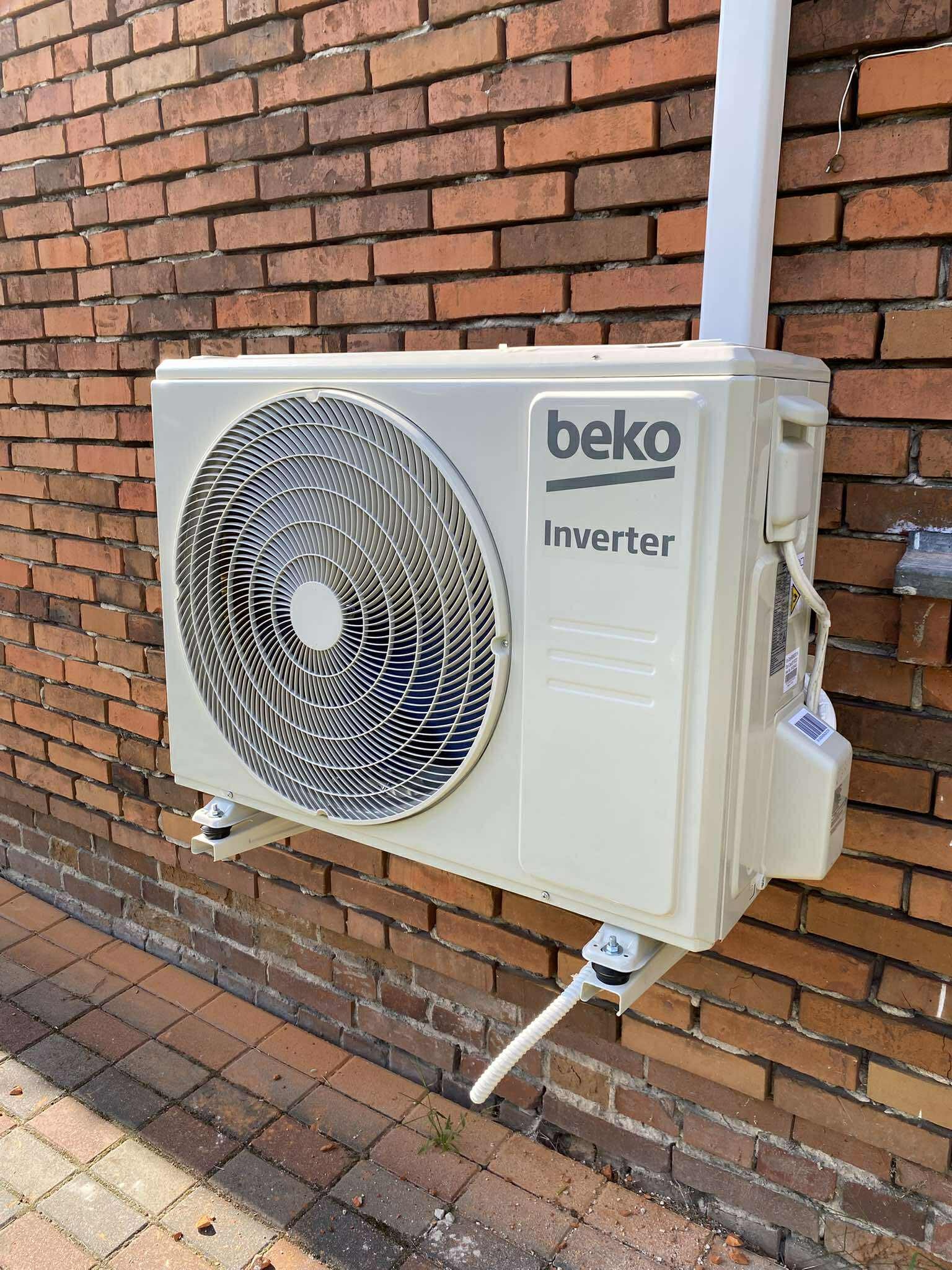 Klimatyzator Beko Inverter zamontowany na ceglanej ścianie zewnętrznej budynku, z widoczną instalacją odprowadzającą skropliny i elementami montażowymi.
