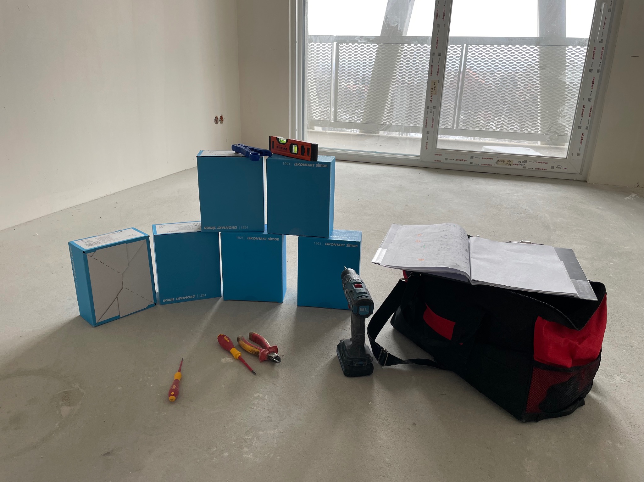 Przygotowanie do montażu osprzętu elektrycznego: puszki, poziomica, wkrętaki, wiertarka i torba narzędziowa na surowym betonie, okno w tle.