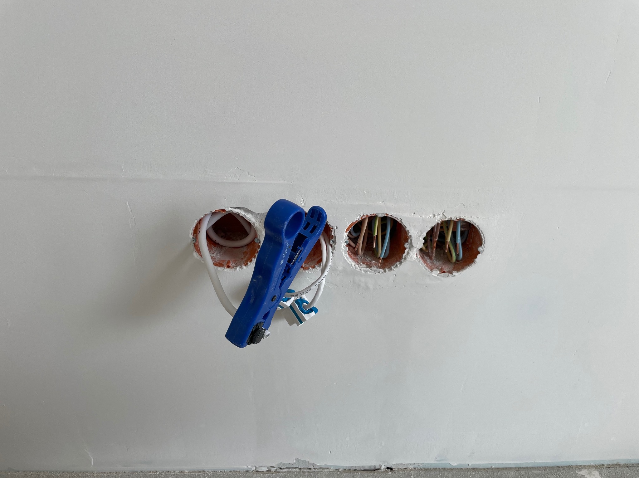 Świeżo położona gładź, trzy otwory w ścianie z przewodami i narzędzie do zdejmowania izolacji. Etap prac elektrycznych w nowym budownictwie.