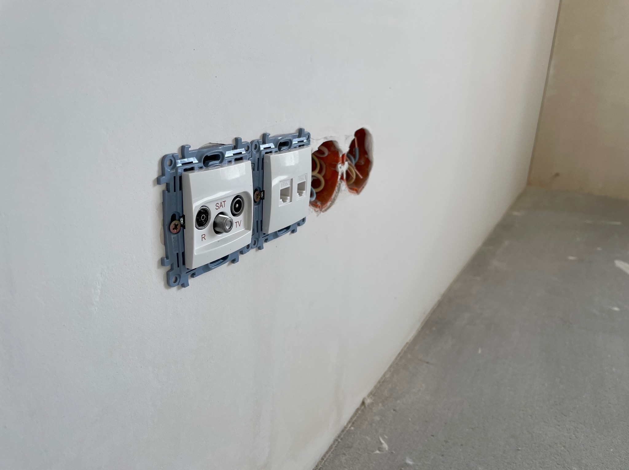 Biała ściana z zamontowanymi gniazdami antenowymi (SAT, TV) i gniazdem sieciowym RJ45. Widoczne przewody w otworach pod gniazda. Szary beton na podłodze.