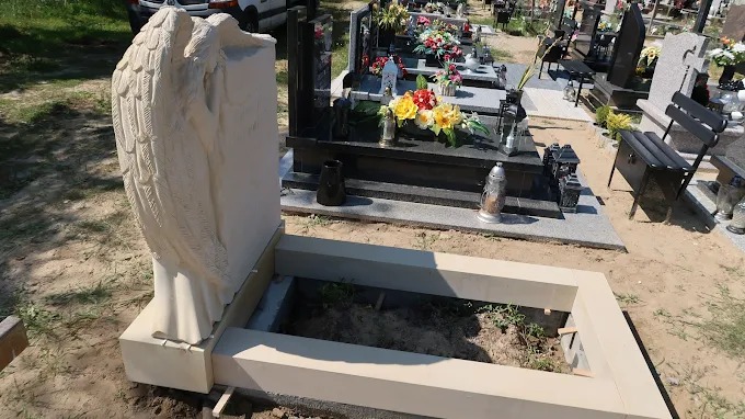 Jasny nagrobek z rzeźbą anioła na cmentarzu, widok z boku. Pusta przestrzeń wewnątrz obramowania nagrobka. Inne nagrobki w tle.