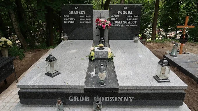 Rodzinny nagrobek z szarego i czarnego kamienia z wygrawerowanymi nazwiskami, ozdobiony kwiatami i lampionami, na tle zieleni cmentarza.