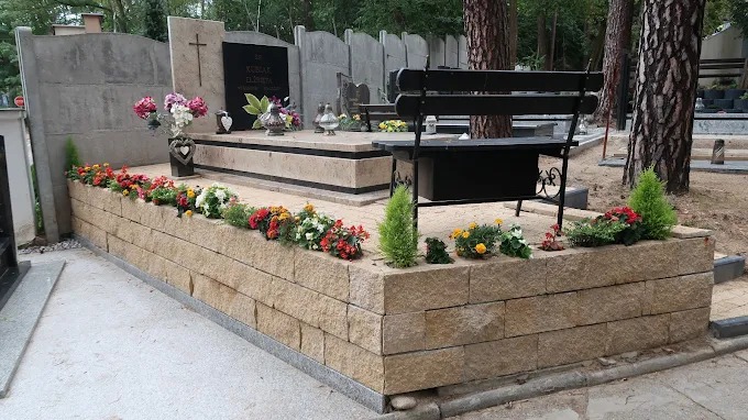 Jasny nagrobek z kamienia naturalnego, otoczony niskim murem z kwiatami i małą ławeczką, na tle drzew na cmentarzu.