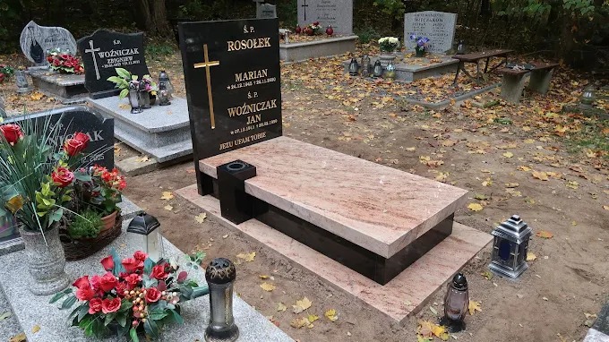 Czarny nagrobek z krzyżem i jasnoróżową płytą poziomą na cmentarzu, otoczony innymi nagrobkami i opadłymi liśćmi. Widok z bliska, jesienna sceneria.