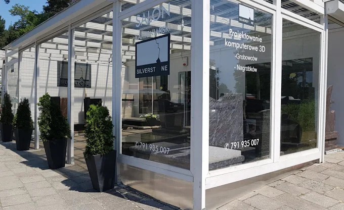Salon kamieniarski Silverstone z ekspozycją nagrobków widoczną przez przeszkloną fasadę. W tle zieleń drzew. Cztery tuje w donicach przed wejściem.