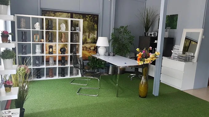 Wnętrze pracowni kamieniarskiej z ekspozycją wazonów nagrobnych na regale, biurkiem i sztuczną trawą na podłodze. Widoczne detale wykończenia i aranżacji.