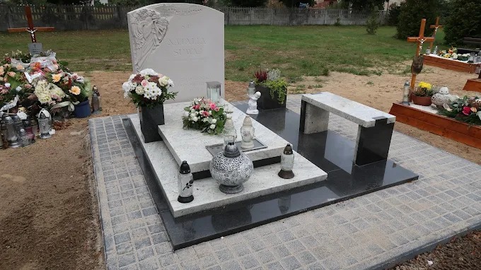 Granitowy nagrobek z rzeźbą anioła, kwiatami i ławką na cmentarzu. Jasne i ciemne kamienie, kostka brukowa wokół. Widok z przodu, w tle zieleń.