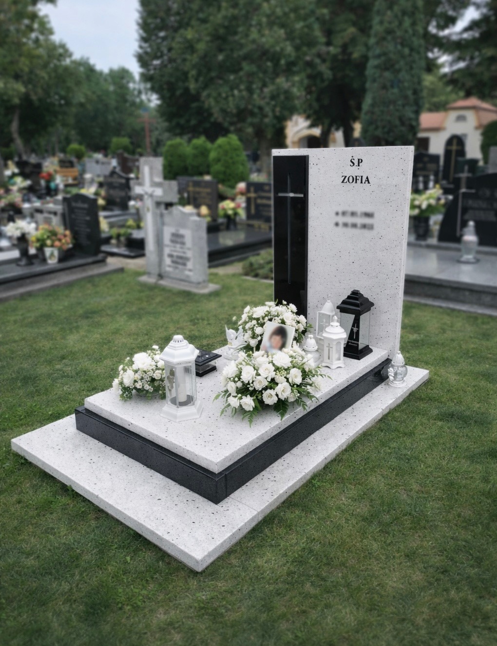 Jasny nagrobek z ciemnym akcentem, ozdobiony białymi kwiatami i lampionami, na tle zielonego trawnika cmentarza. Widoczny portret osoby zmarłej.