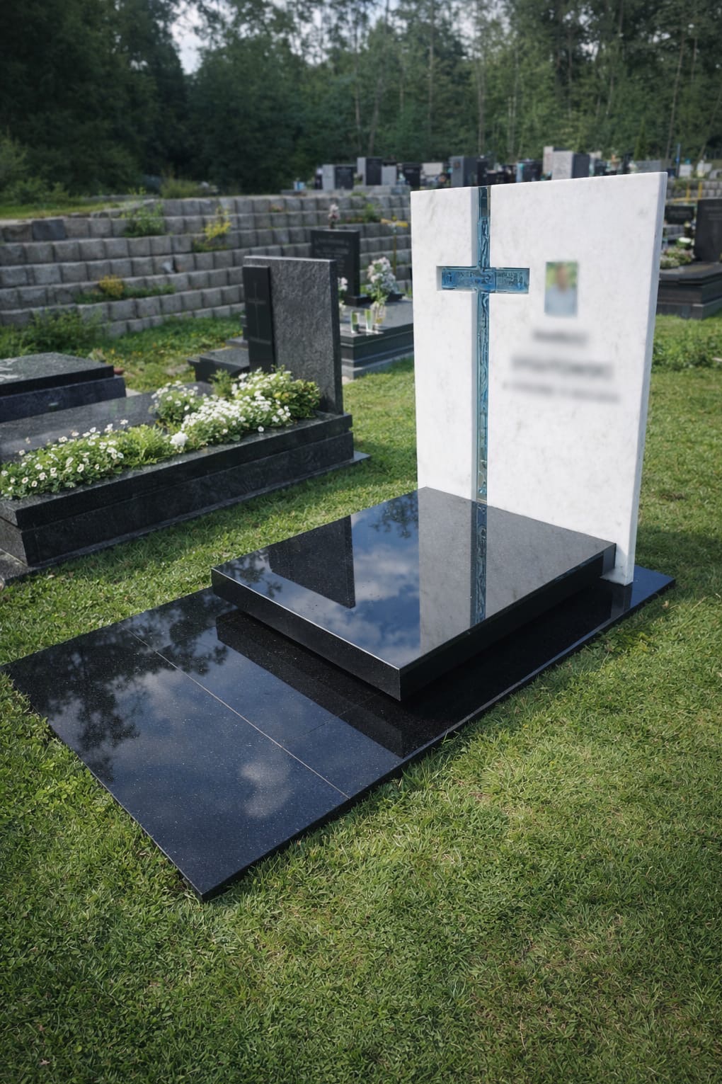 Nowoczesny nagrobek z białego marmuru z niebieskim krzyżem na tle cmentarza, z czarną płytą odbijającą niebo. Starannie utrzymany trawnik.