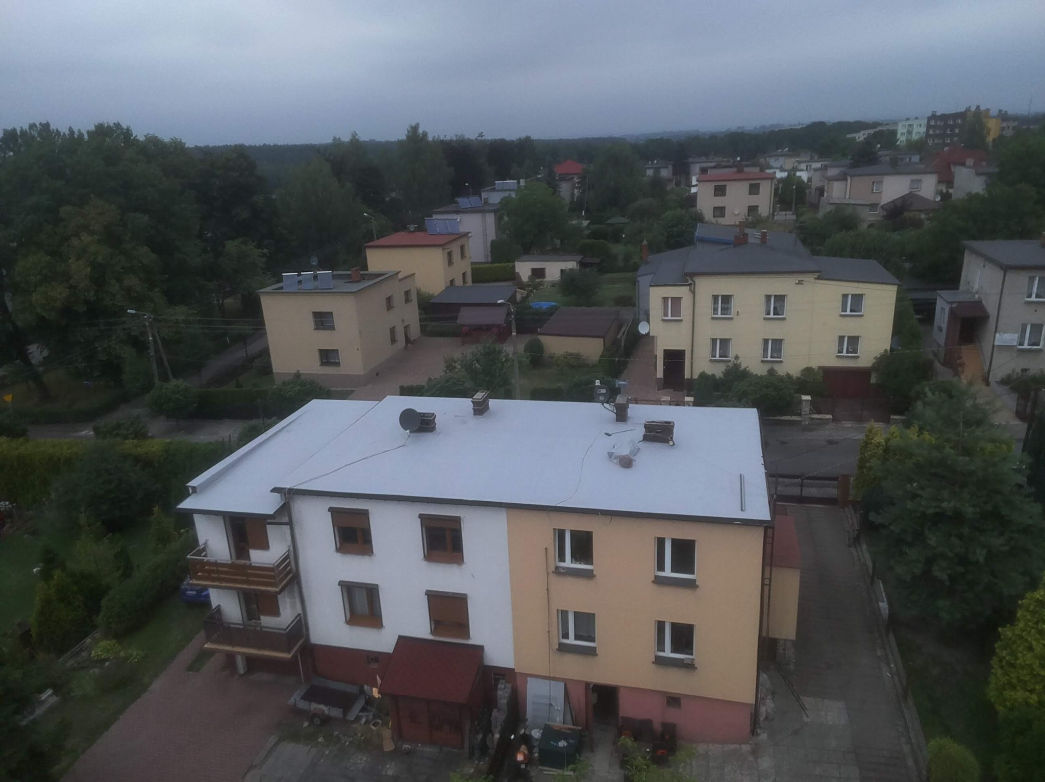 Widok z góry na dwukolorowy budynek z jasnoszarym dachem i kilkoma kominami na tle zabudowy mieszkalnej i drzew w pochmurny dzień.