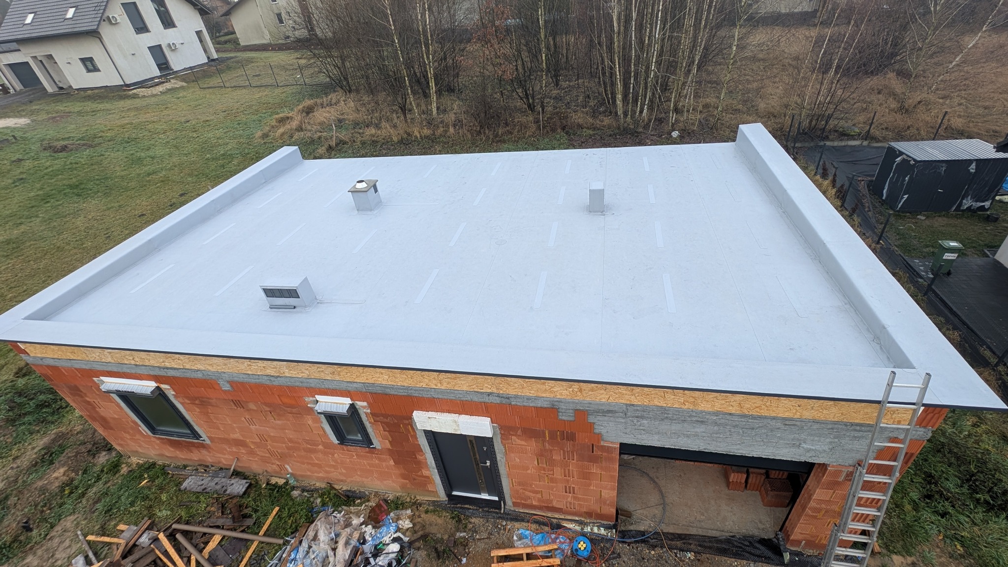 Nowoczesny, płaski dach pokryty jasną membraną, z widocznymi kominkami wentylacyjnymi i detalami wykończeniowymi. Budynek w trakcie budowy, z widoczną konstrukcją ścian.