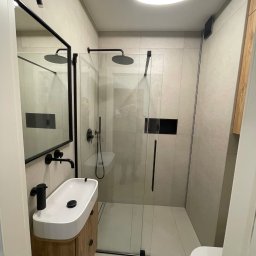 MIK-BUD Mikołaj Patrzała - Nowoczesna łazienka z kabiną prysznicową typu walk-in, umywalką nablatową na drewnianej szafce i czarną armaturą. Minimalistyczny design, jasne płytki i oświetlenie LED.