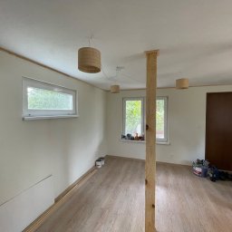 MIK-BUD Mikołaj Patrzała - Jasne wnętrze po remoncie: świeżo pomalowane ściany, drewniana podłoga, okna z widokiem na zieleń, lampy w stylu boho i drewniany słup konstrukcyjny.