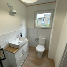 MIK-BUD Mikołaj Patrzała - Jasna łazienka z umywalką na szafce z drewnianym blatem, toaletą i oknem z widokiem na budynek. Ściany wyłożone białymi płytkami, podłoga drewnopodobna.