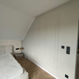 MIK-BUD Mikołaj Patrzała - Jasne wnętrze poddasza z łóżkiem i panelami ściennymi. Parkiet w jodełkę, minimalistyczna lampka nocna. Widoczny fragment osoby w przejściu do innego pomieszczenia.