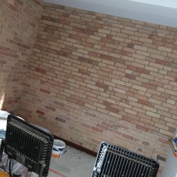 MIK-BUD Mikołaj Patrzała - Ściana wykończona cegłą klinkierową w odcieniach beżu i brązu, oświetlona dwoma halogenami budowlanymi. Widoczny fragment sufitu i podłogi w trakcie prac wykończeniowych.