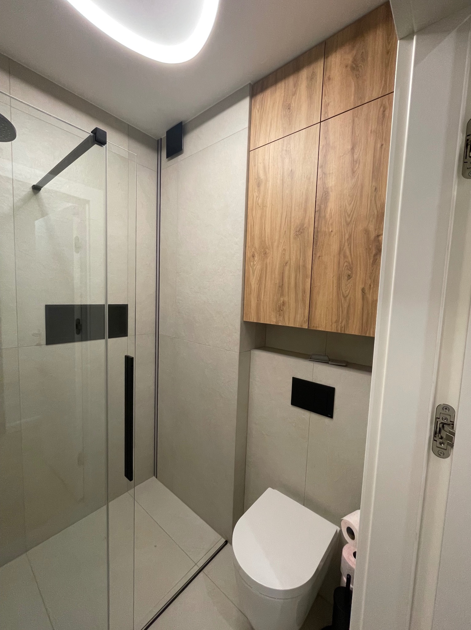 Nowoczesna łazienka po remoncie: kabina prysznicowa typu walk-in z czarnymi detalami, podwieszana toaleta i szafka z drewnianym frontem. Minimalistyczny design, jasne płytki.