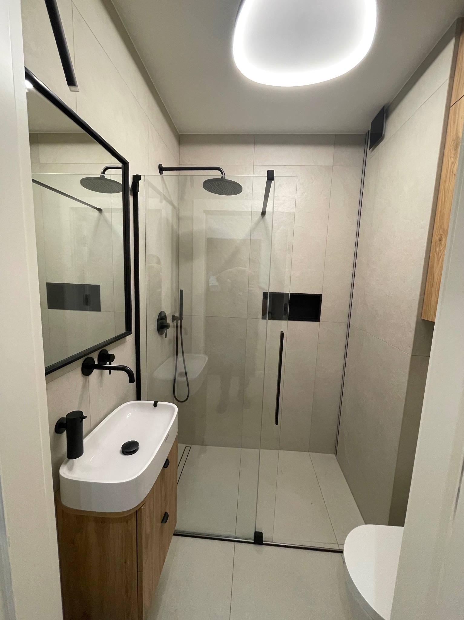 Nowoczesna łazienka z kabiną prysznicową typu walk-in, umywalką nablatową na drewnianej szafce i czarną armaturą. Minimalistyczny design, jasne płytki i oświetlenie LED.