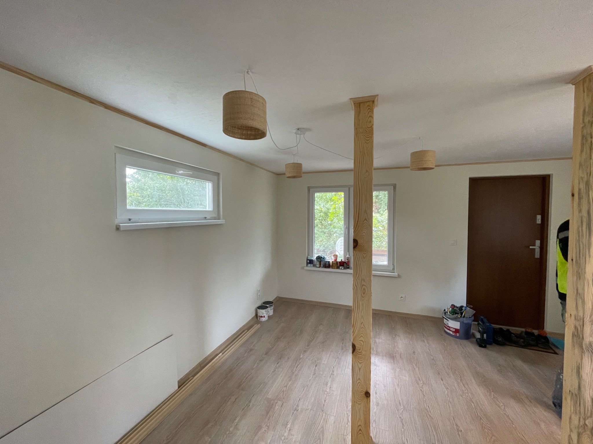 Jasne wnętrze po remoncie: świeżo pomalowane ściany, drewniana podłoga, okna z widokiem na zieleń, lampy w stylu boho i drewniany słup konstrukcyjny.