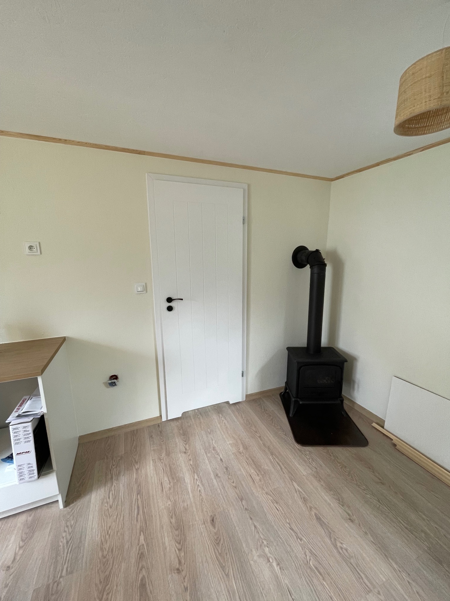 Jasne wnętrze z panelami podłogowymi, białymi ścianami z listwami, białymi drzwiami i czarnym piecem wolnostojącym. Widoczny fragment szafki i lampy.