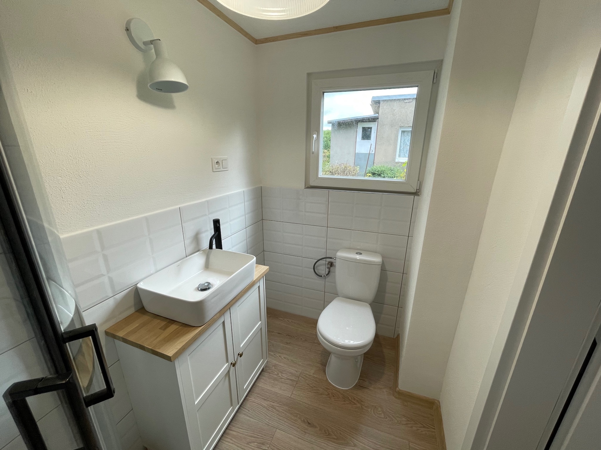Jasna łazienka z umywalką na szafce z drewnianym blatem, toaletą i oknem z widokiem na budynek. Ściany wyłożone białymi płytkami, podłoga drewnopodobna.