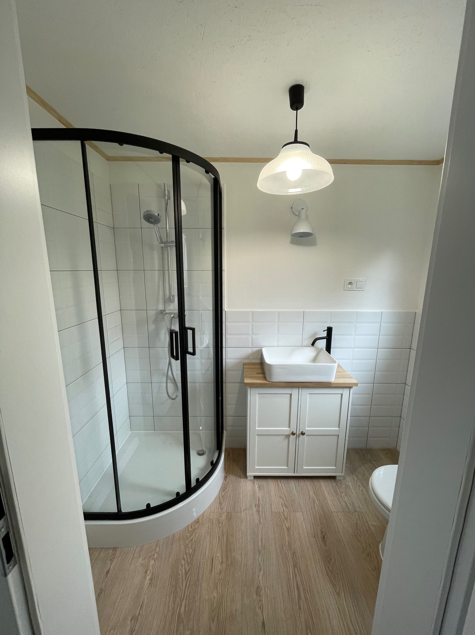 Nowoczesna łazienka po remoncie: kabina prysznicowa z czarnymi ramami, biała umywalka na szafce z drewnianym blatem, białe płytki na ścianach i drewnopodobna podłoga.