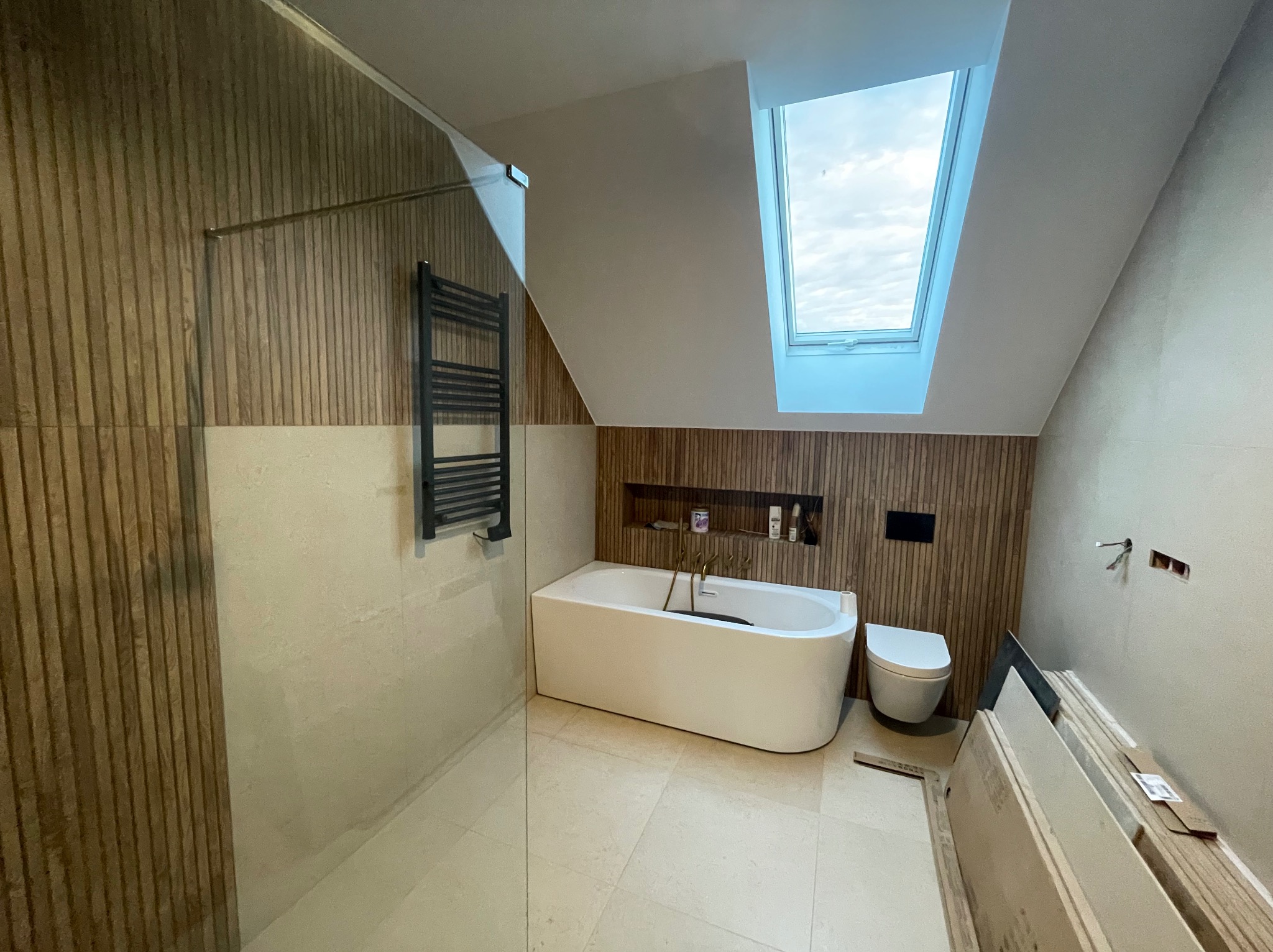 Nowoczesna łazienka na poddaszu z drewnianą okładziną, białą wanną i oknem dachowym. Minimalistyczny design, jasne płytki podłogowe, grzejnik drabinkowy.