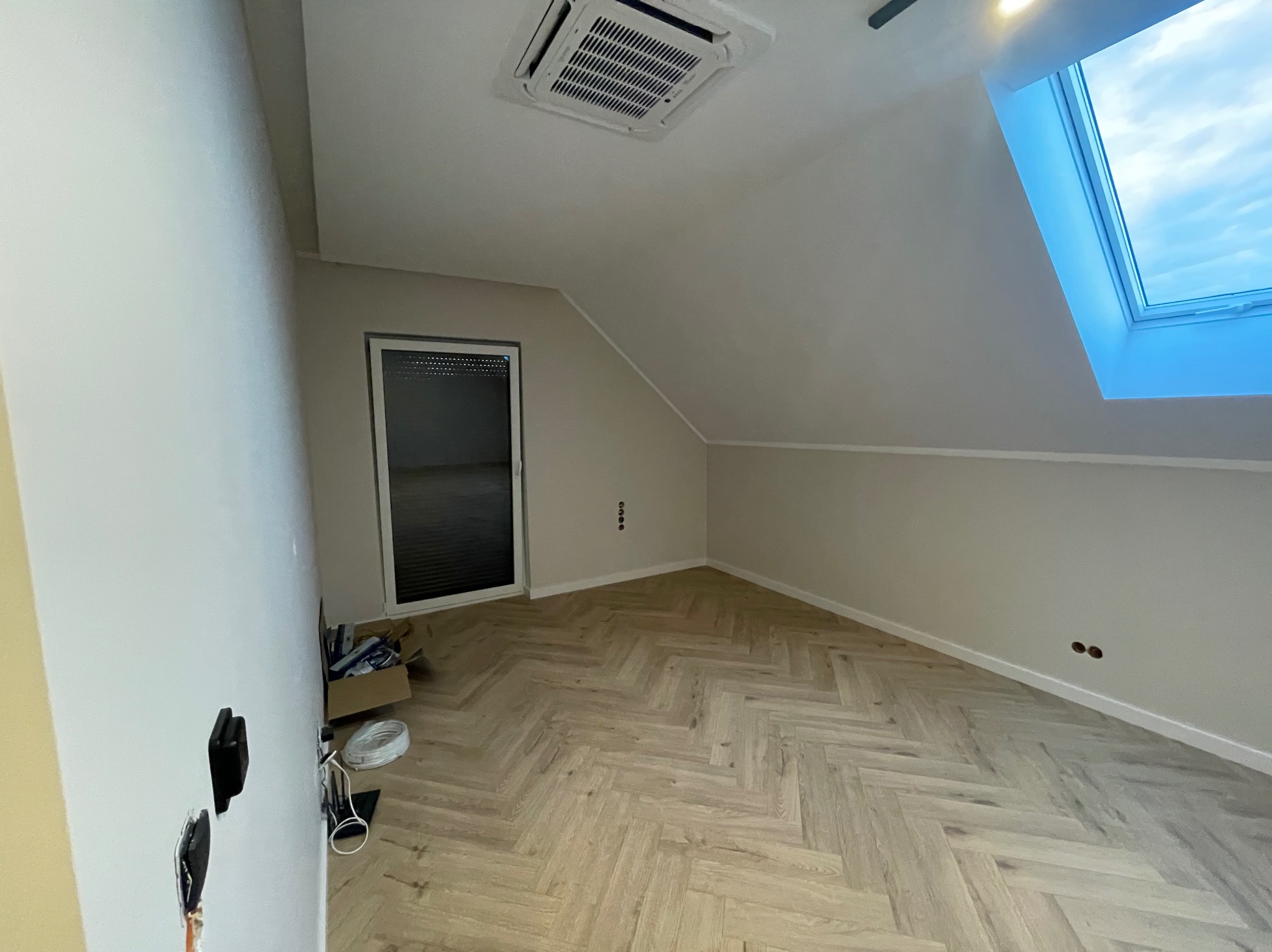 Jasne wnętrze poddasza z parkietem w jodełkę, oknem dachowym i klimatyzacją sufitową. Widoczne przygotowania do montażu elektryki.