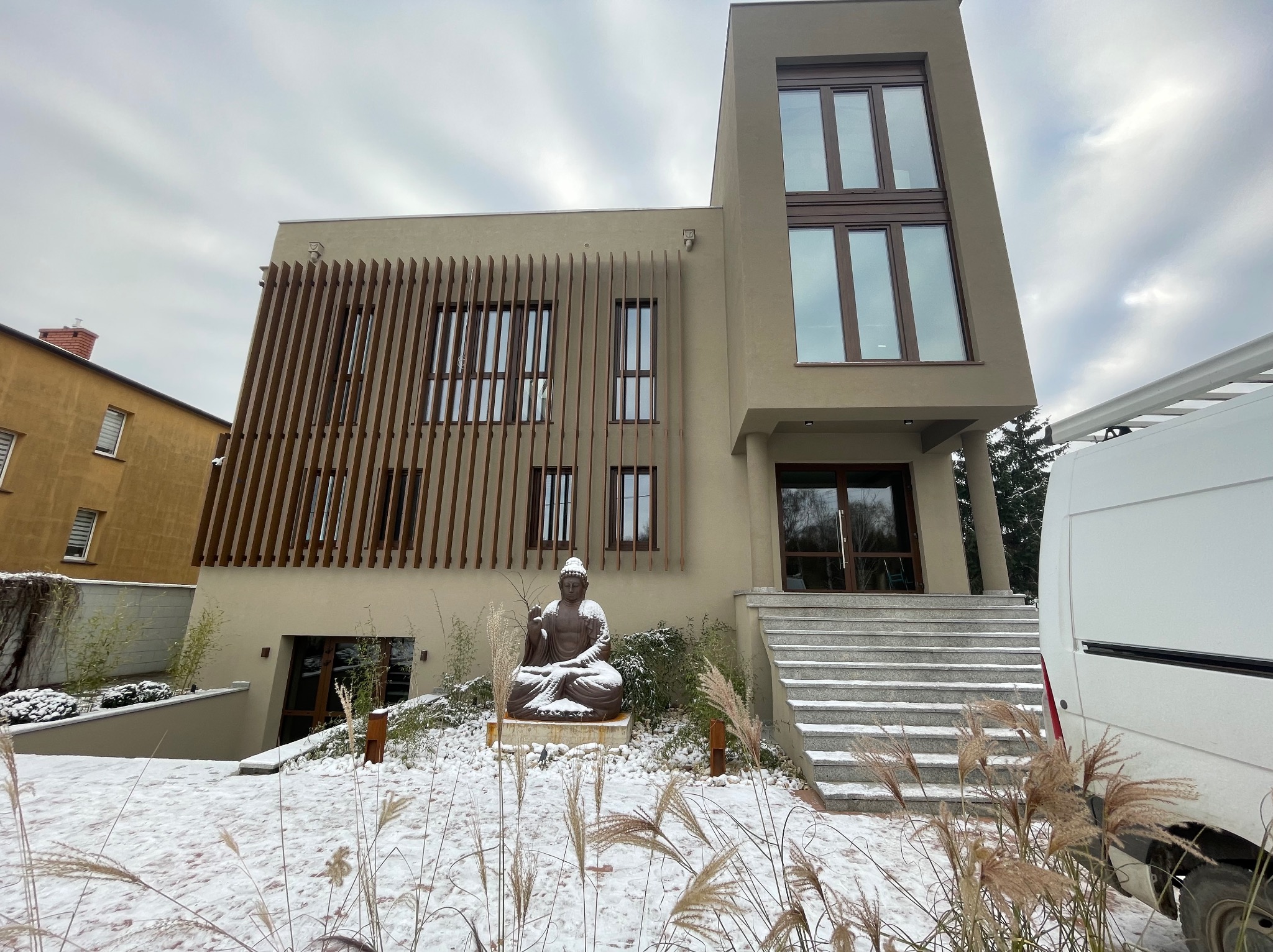 Nowoczesny budynek z drewnianą fasadą i rzeźbą Buddy w zimowej scenerii. Widoczny fragment białego samochodu dostawczego.