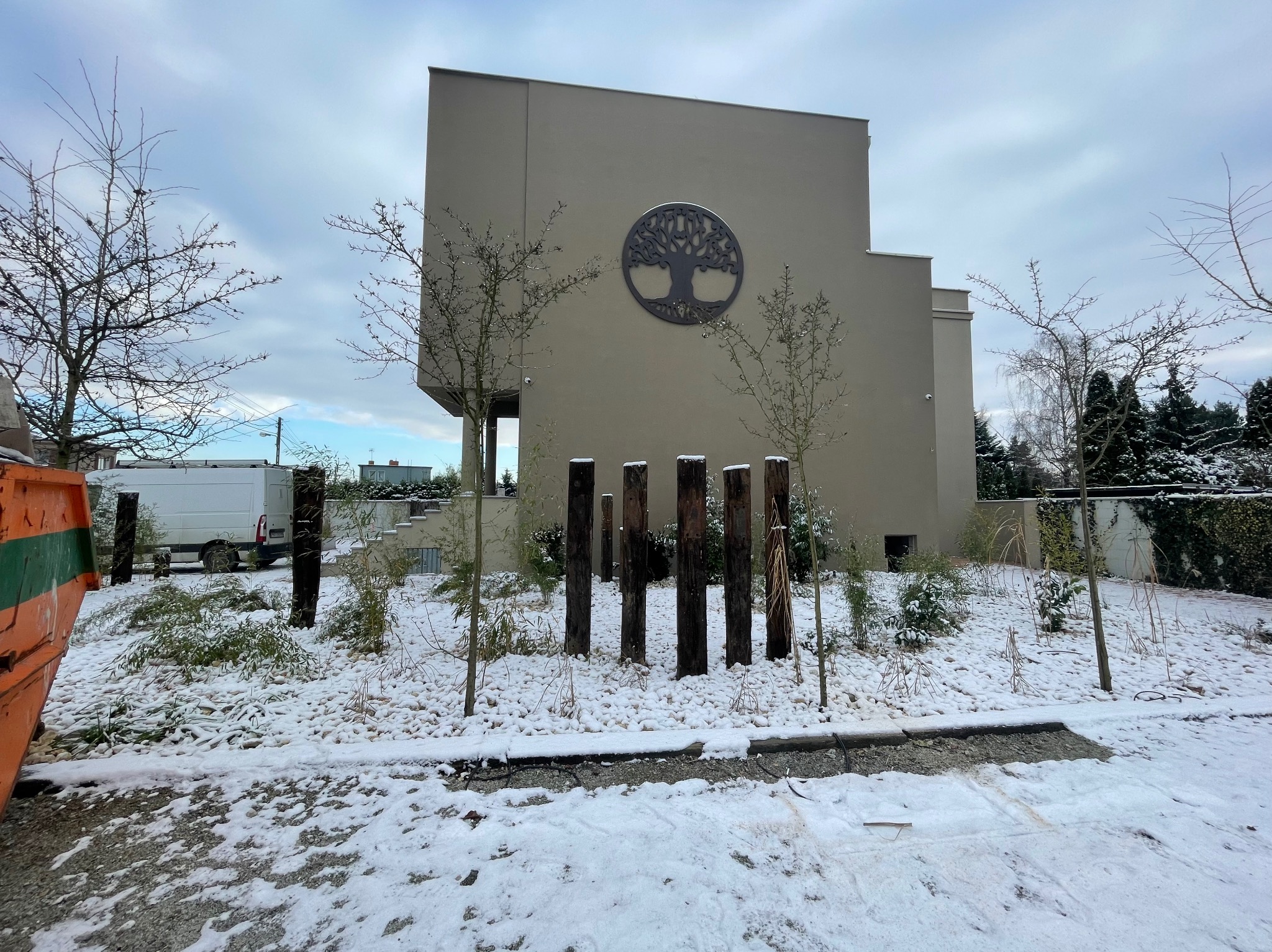 Nowoczesny dom z beżową elewacją i dekoracyjnym drzewem na ścianie. Ogród w zimowej scenerii, z drewnianymi elementami i śniegiem.