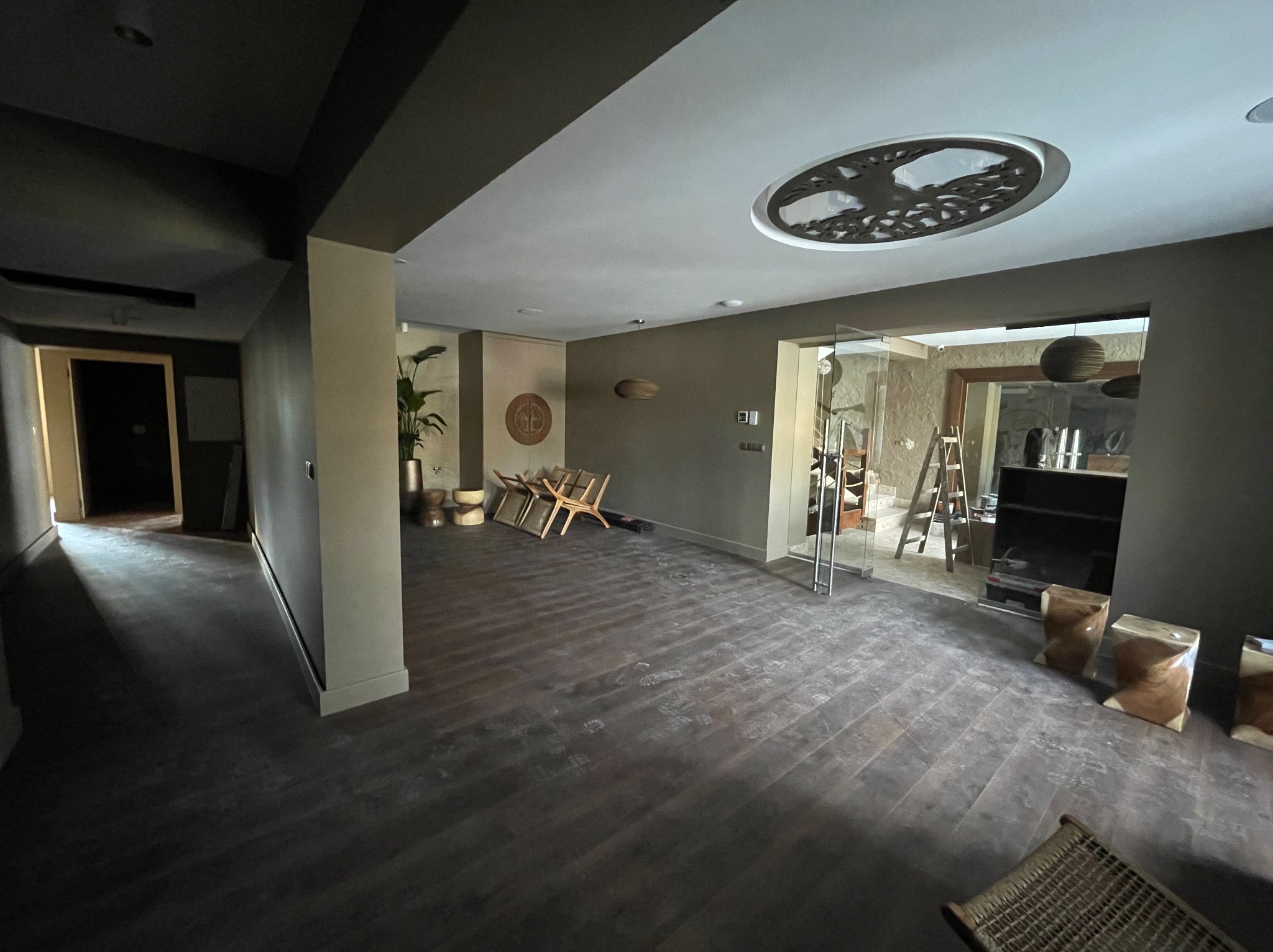 Przestronne wnętrze z ciemną podłogą ułożoną w jodełkę, dekoracyjnym oświetleniem sufitowym i elementami wystroju w stylu boho. Widoczne krzesła i rośliny doniczkowe.