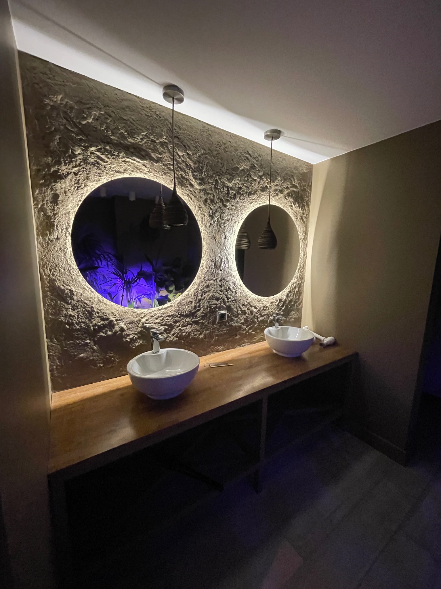 Nowoczesna łazienka z dwoma okrągłymi lustrami z podświetleniem LED, umywalkami nablatowymi i blatem z drewna. Ściana z fakturą, wiszące lampy.