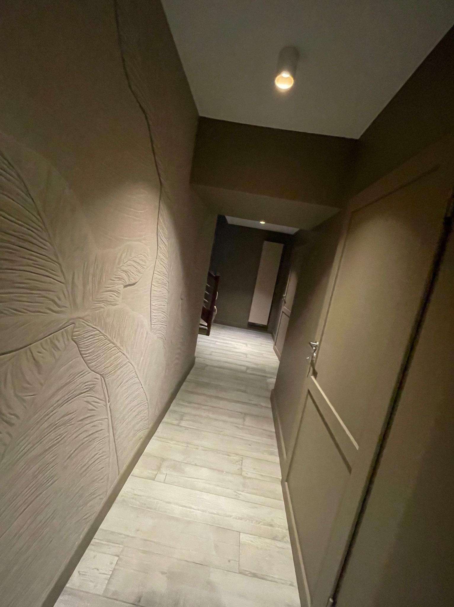 Korytarz w odcieniach brązu z dekoracyjną ścianą z motywem liści, jasną podłogą i oświetleniem punktowym. Widoczne drzwi i fragment dalszej części korytarza.