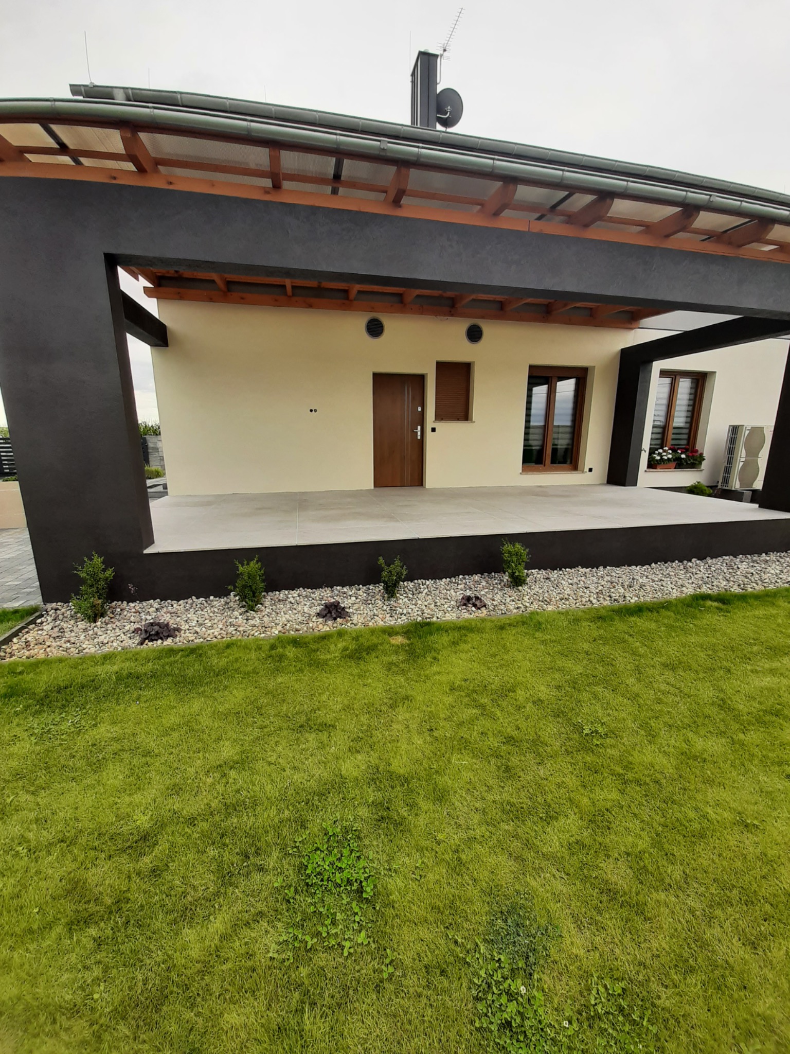 Nowoczesny dom jednorodzinny z zadaszonym tarasem, beżową elewacją i ciemnymi elementami konstrukcyjnymi. Widoczny trawnik i ozdobne kamienie.