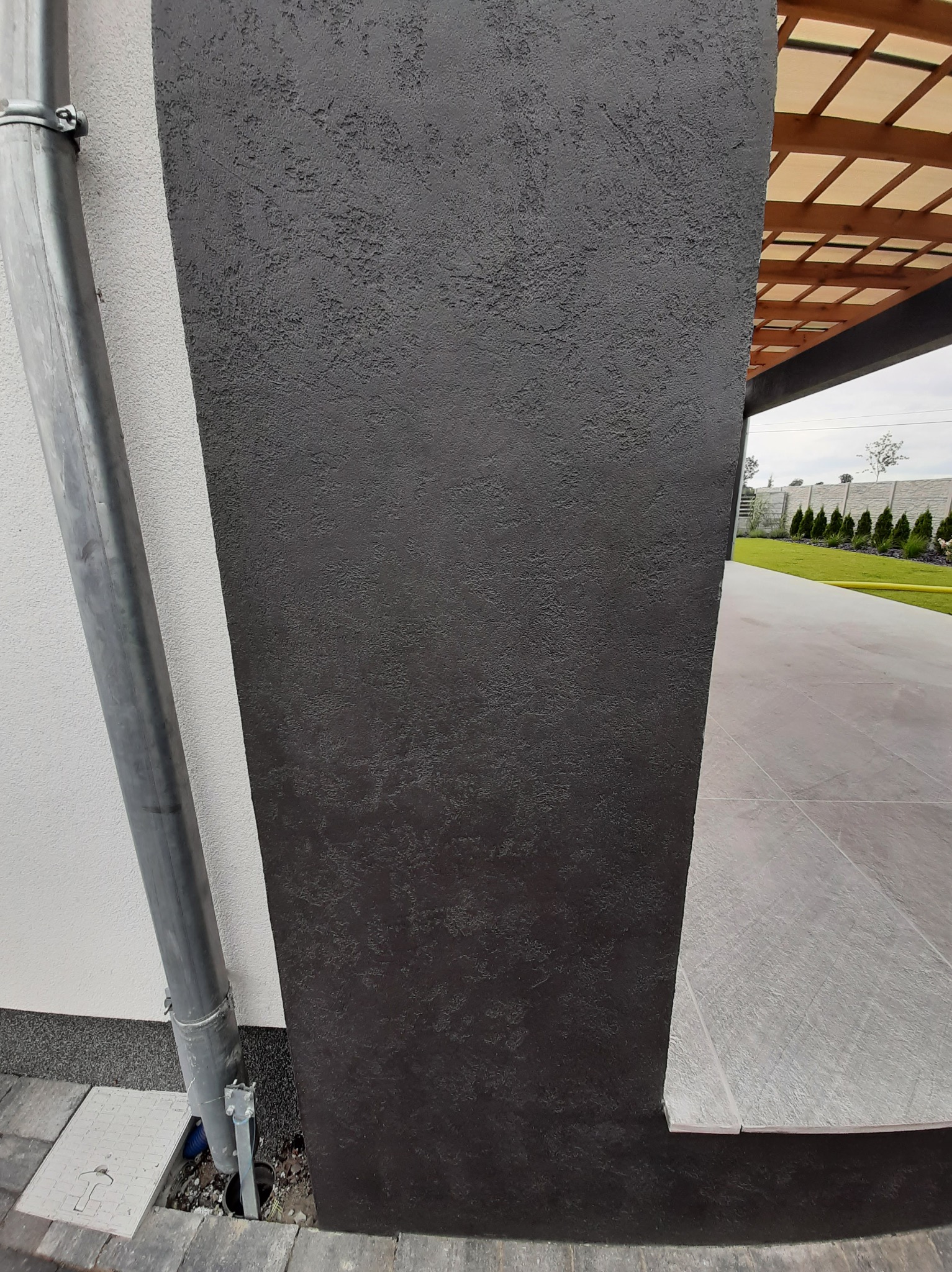 Czarny, teksturowany filar z rynną na tle białej ściany i szarej posadzki. Widoczny fragment drewnianej konstrukcji zadaszenia i zielony trawnik w tle.