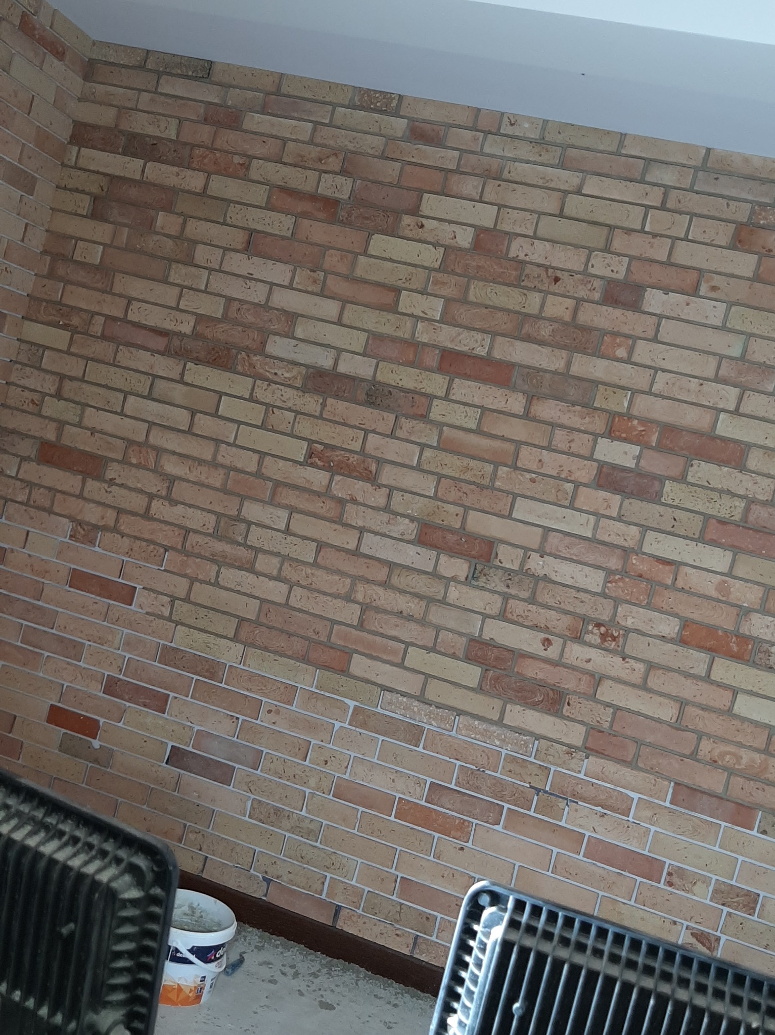 Ściana z cegły klinkierowej w odcieniach beżu i brązu, widoczny fragment grzejnika i wiaderko z farbą, sugerujące prace wykończeniowe.