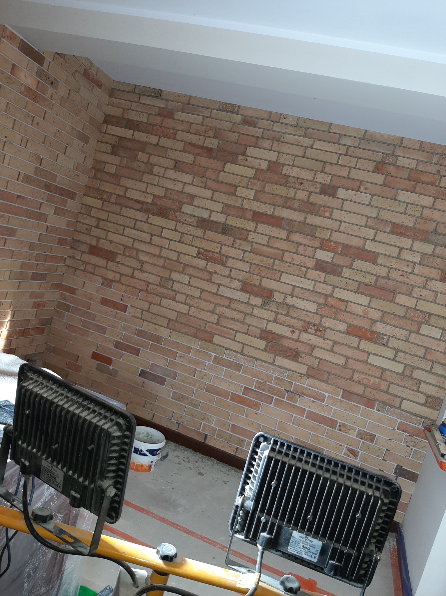 Ściana wykończona cegłą klinkierową w odcieniach beżu i brązu, oświetlona dwoma halogenami budowlanymi. Widoczny fragment sufitu i podłogi w trakcie prac wykończeniowych.