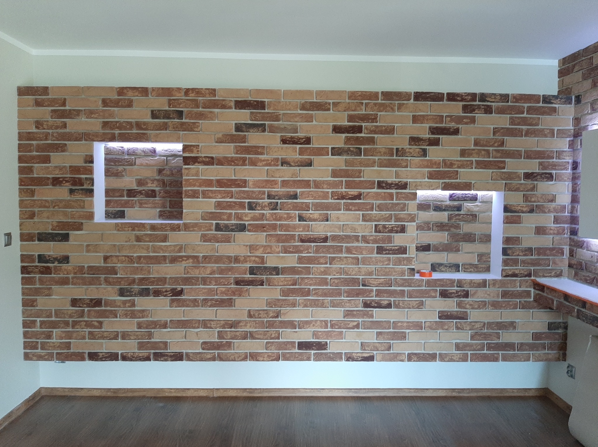 Ściana z cegły dekoracyjnej z dwoma podświetlanymi wnękami. Widoczna podłoga z paneli i fragment blatu. Ciepłe oświetlenie wnęk.